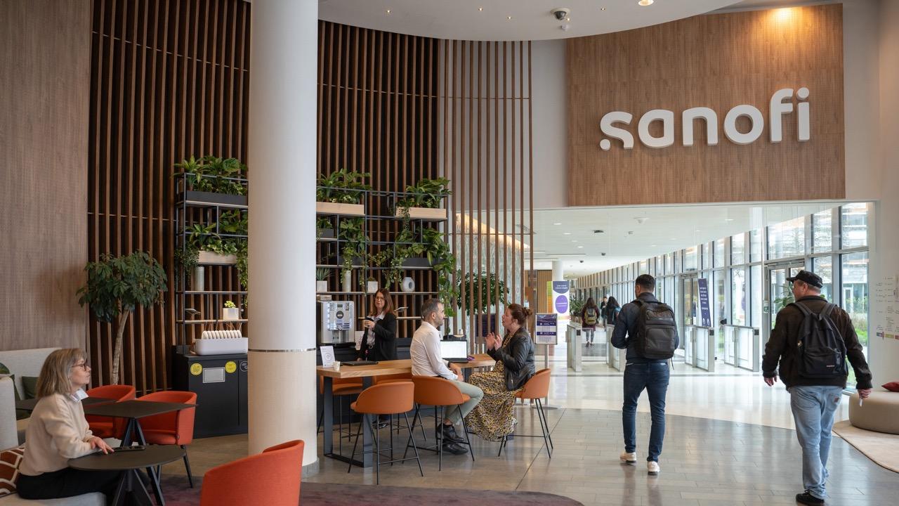 Sanofi office entrance area featuring modern design with wooden accents, glass walls, and greenery. The Sanofi logo is displayed on a large wooden sign. Several people are present - seated at orange chairs, working, or conversing - in a bright, welcoming common space.