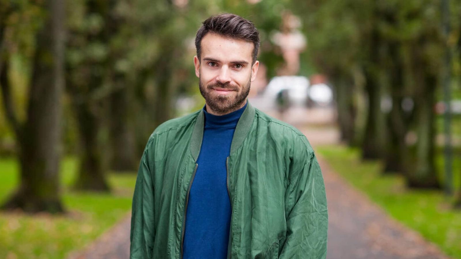 Vegard, paciente de dermatitis atópica, mira fijamente a cámara en un parque con jardines. Viste chaqueta verde y camiseta azul.