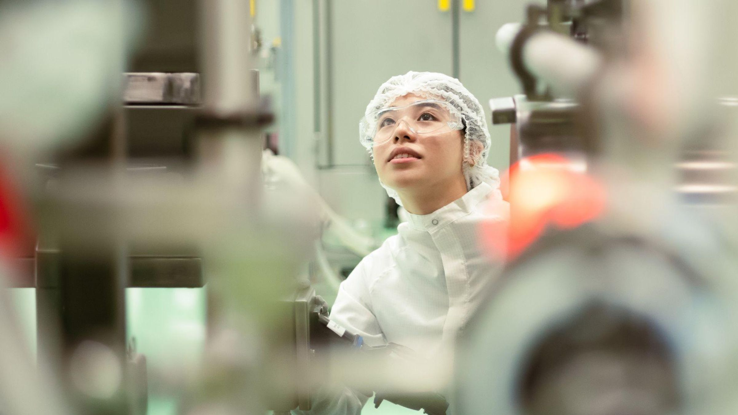 Woman wearing a white sterile suit and hair cover, operating equipment in a pharmaceutical production area.
