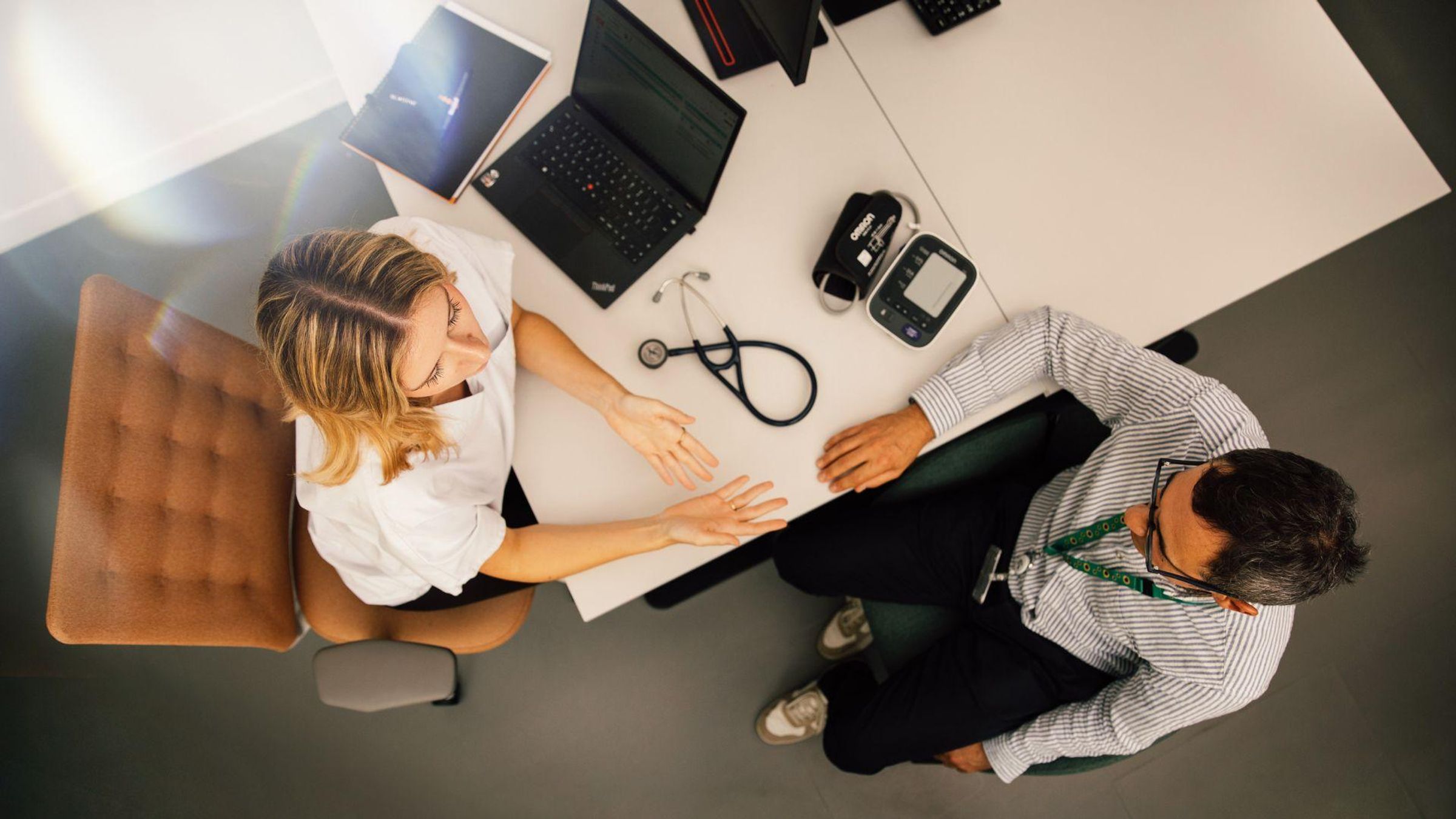 Overhead view of a doctor in white and patient in striped shirt in consultation, with medical equipment including a stethoscope and blood pressure monitor on the desk between them