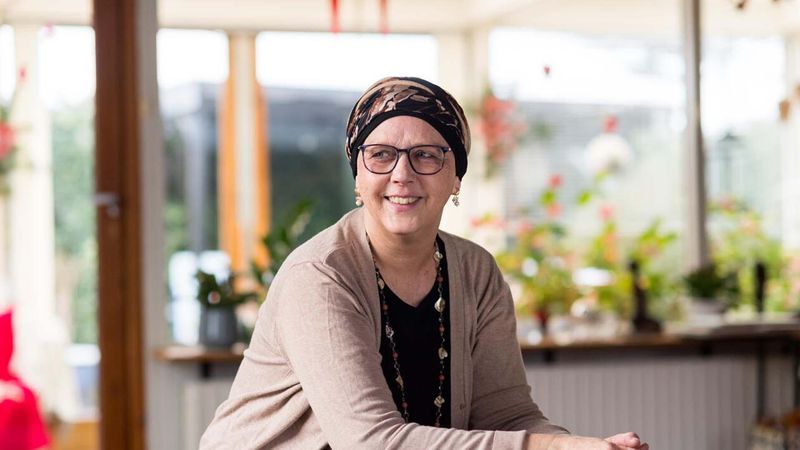 A woman sitting indoors, wearing a beige cardigan and head scarf, smiling at the camera. Behind her are a glass door, shelves with plants, and a window