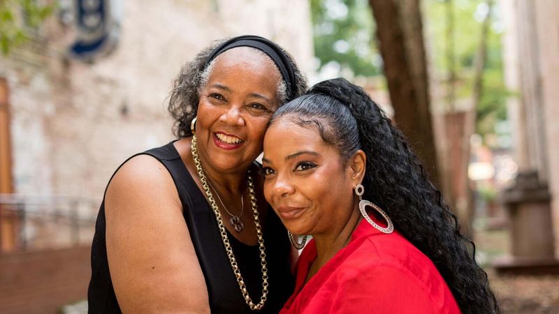 Two smiling women standing outdoors. One wears a black sleeveless top, the other a red top with hoop earrings. Trees and a building are visible in the background