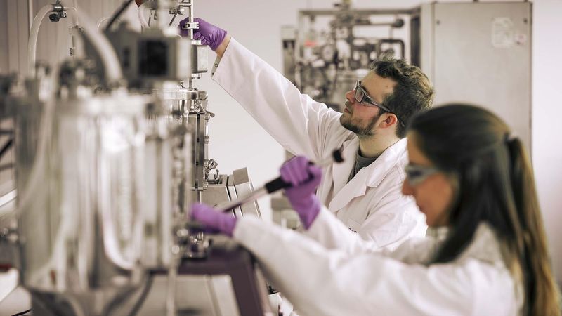 Two scientists, a man and a woman, working in a laboratory. Both wear white lab coats. The man has a bandana and glasses, the woman wears gloves. Laboratory equipment is visible in the background.