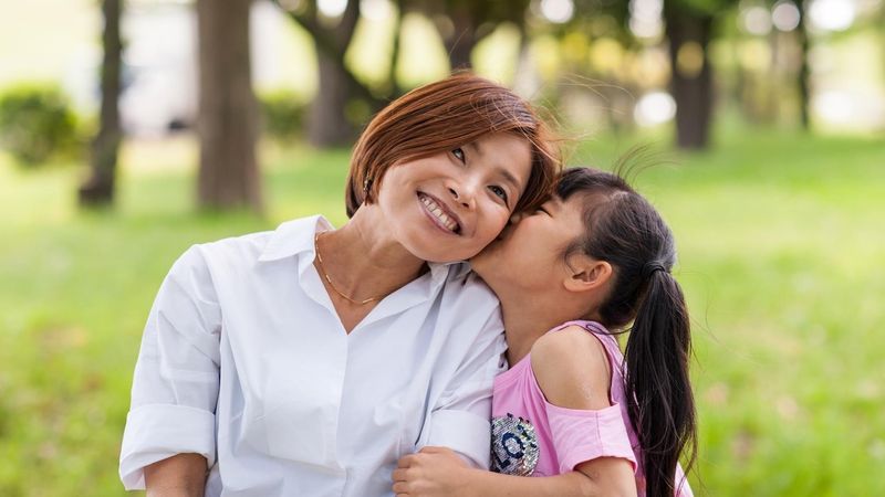 Miwako a woman living with an immunology disease smiling at the camera while her daughter kisses her on the cheek in a park-like setting.