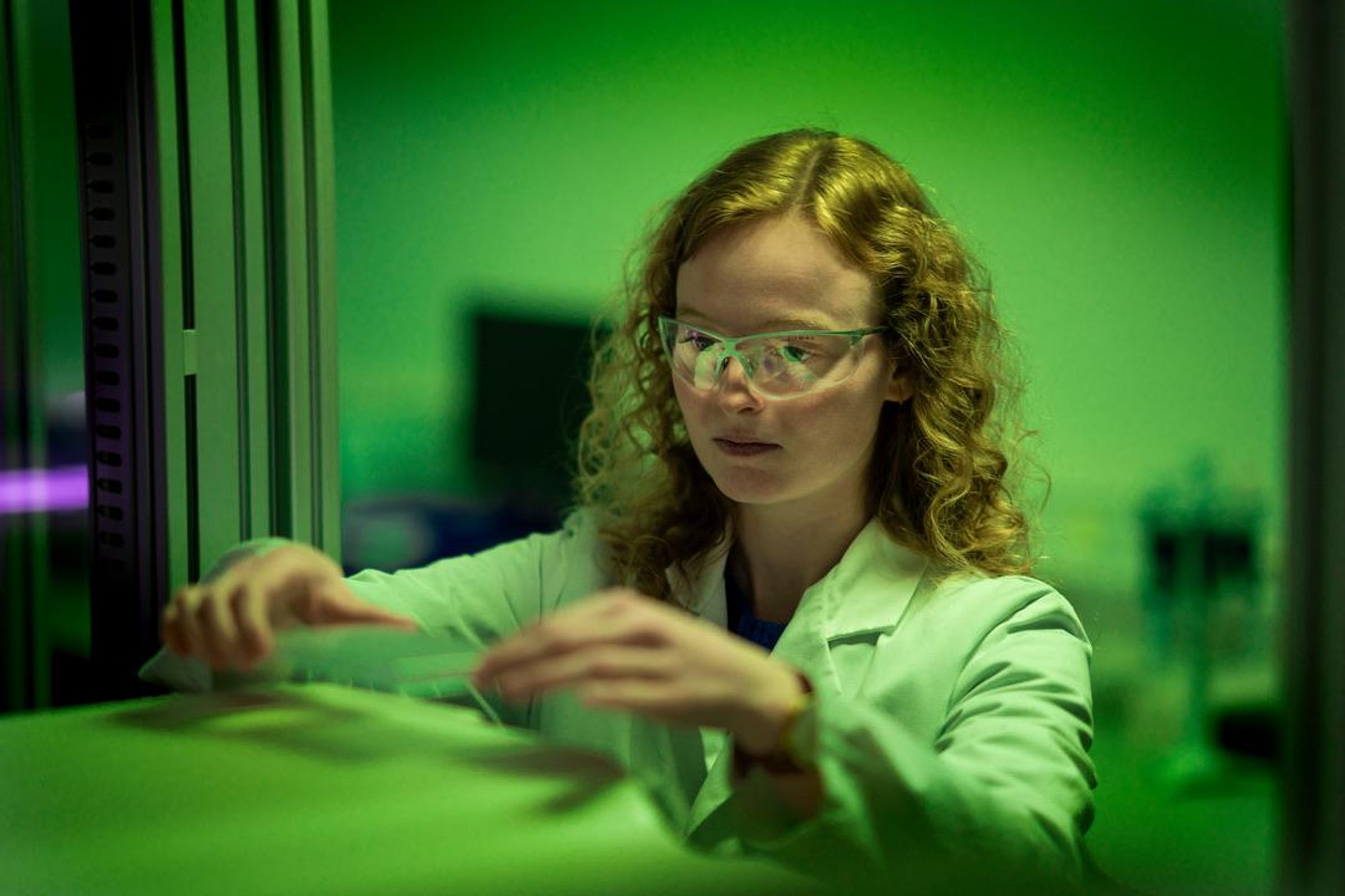 Camille De Craene has long curly blond hair, wearing safety glasses and a white lab coat, and holding glass in a green lit room