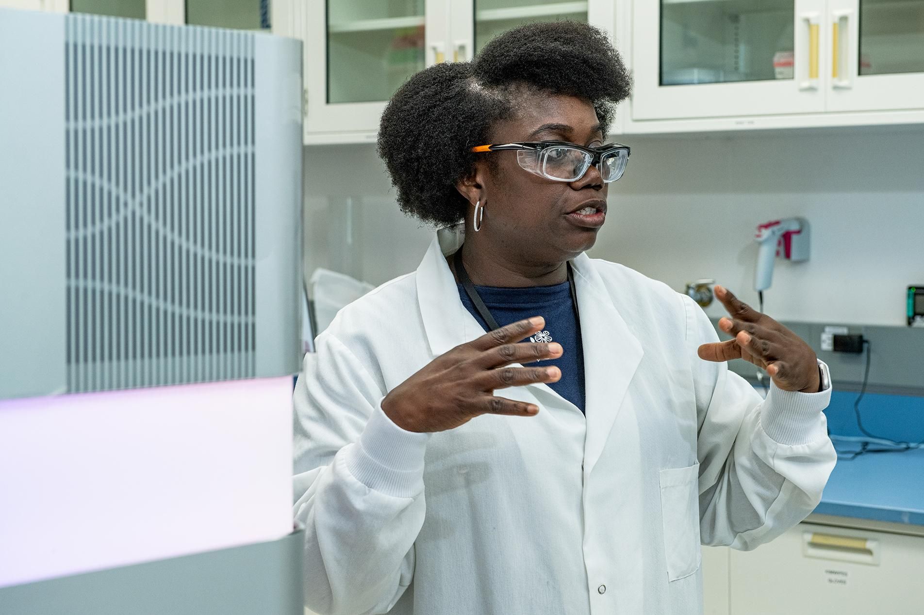 Francisca Aidoo-Nyarko, wearing a white lab coat, safety glasses and hoop earrings, makes hand gestures as she speaks