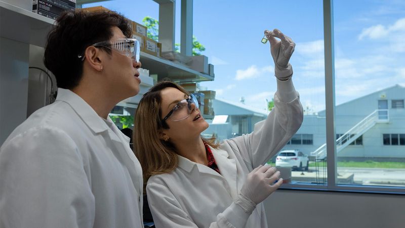 Sung-In Back and Sahar Esmaeili Samani, both wearing white lab coats and safety glasses, examine a partially filled vial