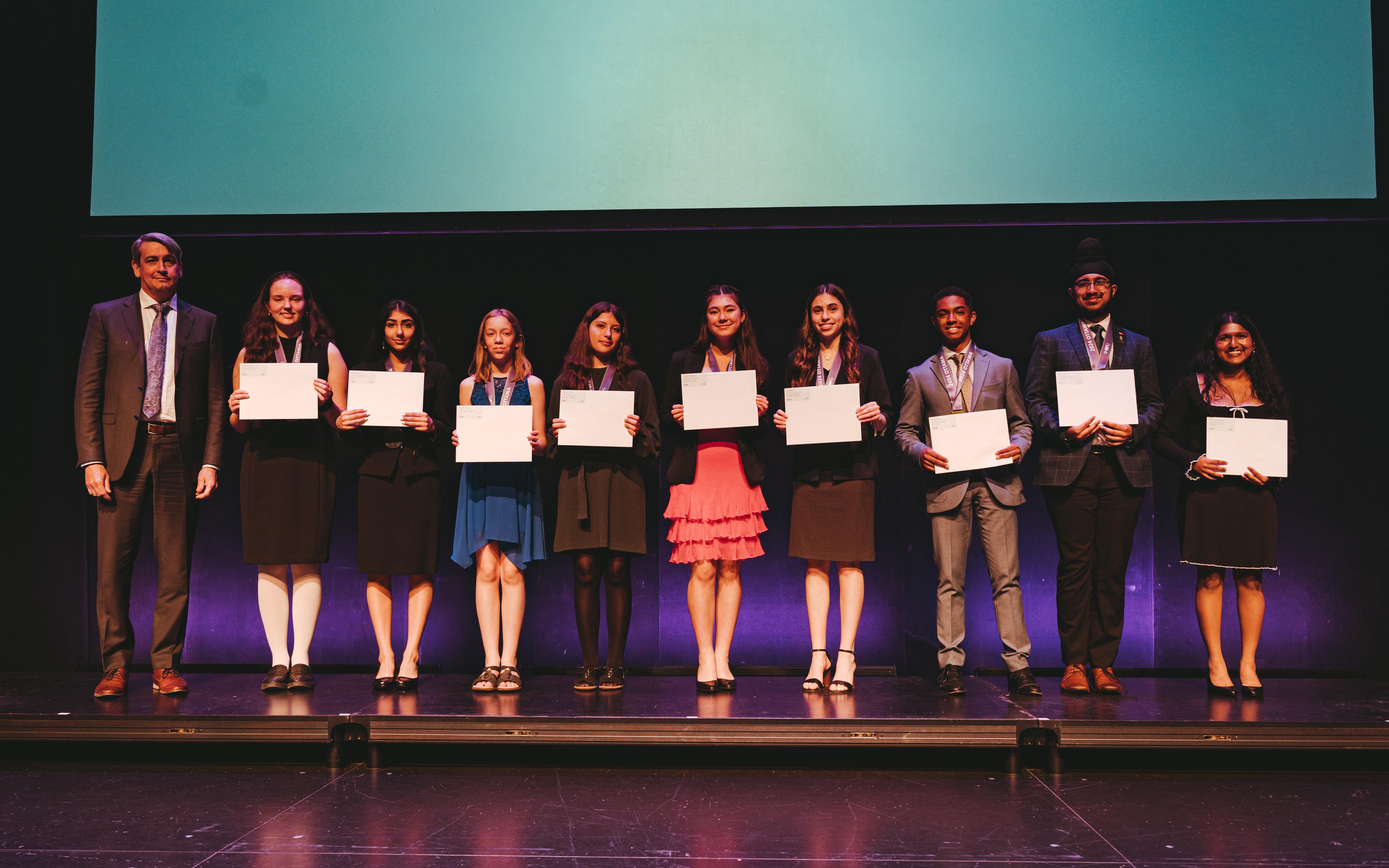 The nine 2024 Biogenius Competition winners, smiling, pose with their certificates on a stage next to a man in a suit