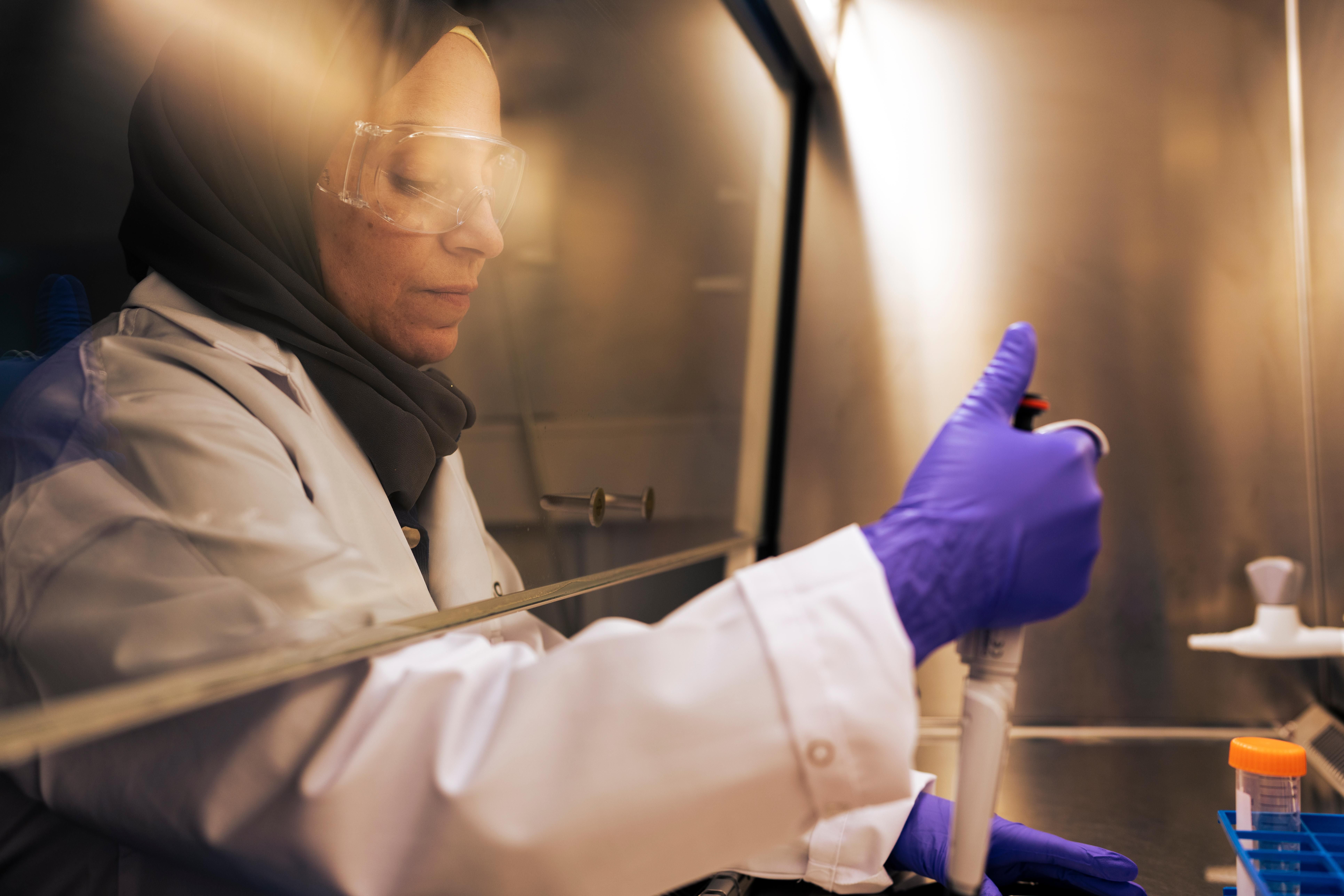 Nadin Al Shukor prepares samples under a fume hood, wearing safety glasses, purple gloves, a white lab coat, and a head scarf