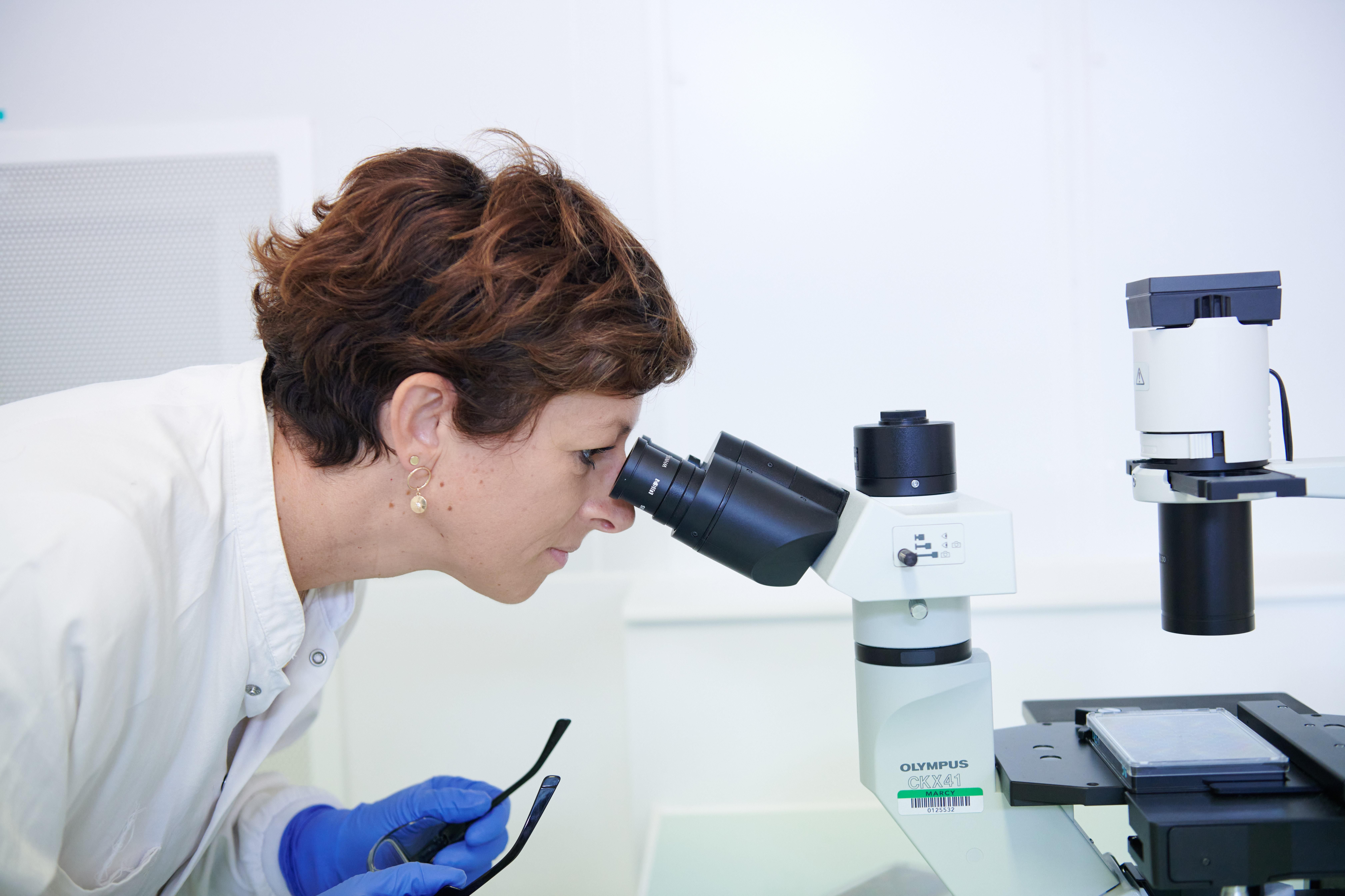 Emilie Proust, with short brown hair and a white lab coat, takes her glasses off to look in the microscope