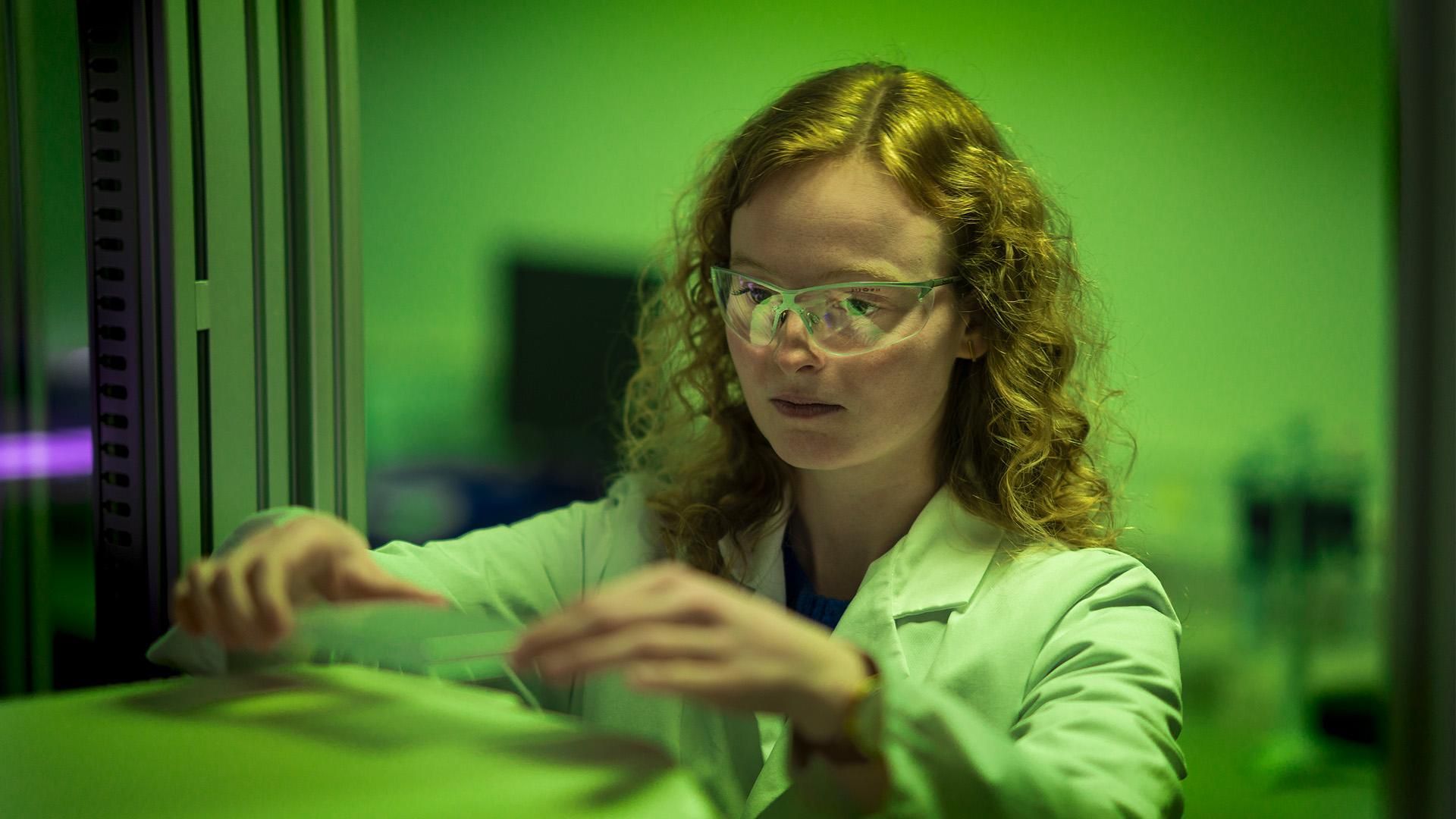 Camille De Craene has long curly blond hair, wearing safety glasses and a white lab coat, and holding glass in a green lit room