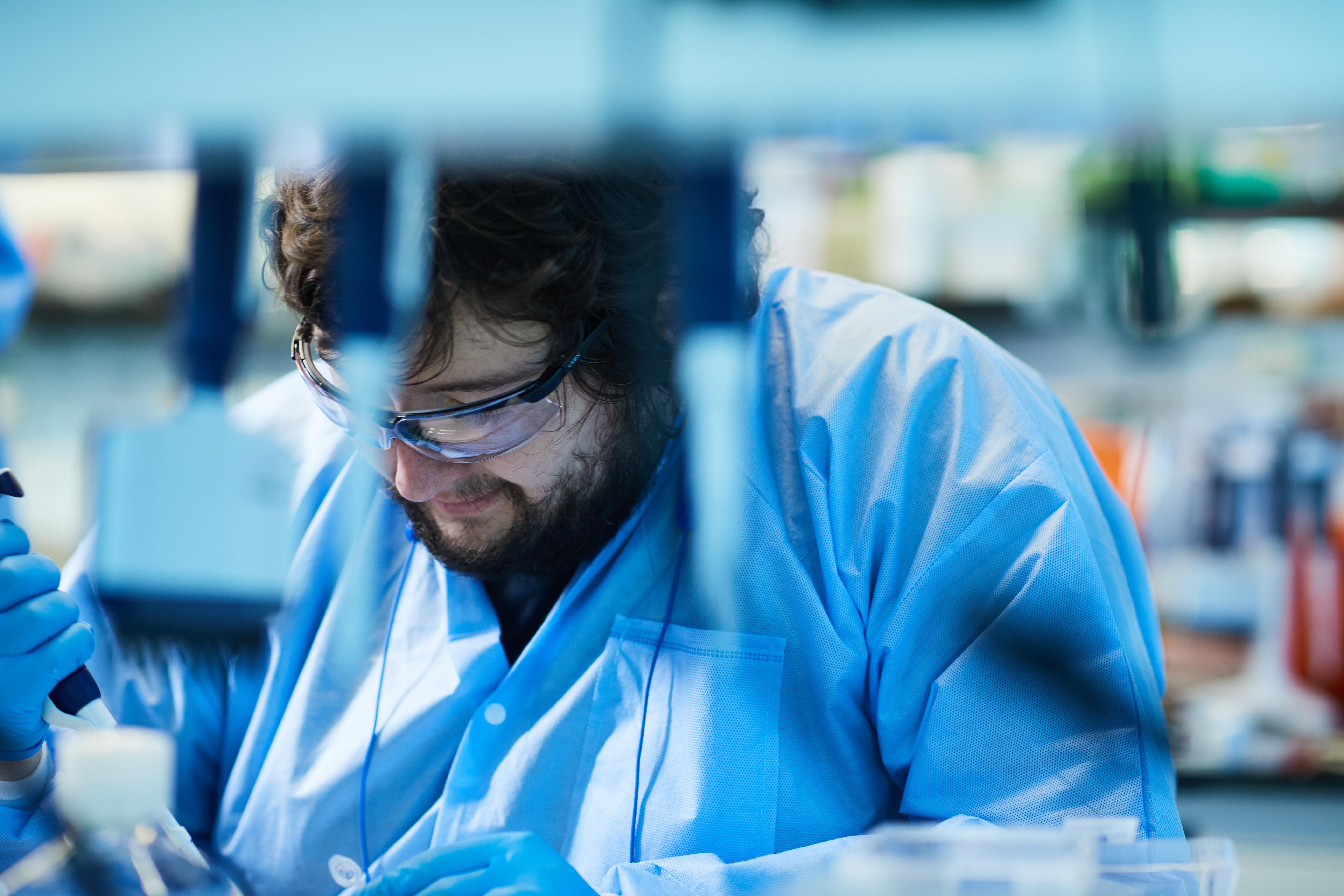 Nick Clark, with curly dark hair and beard, wearing blue gloves, lab outfit, and safety glasses, working with samples