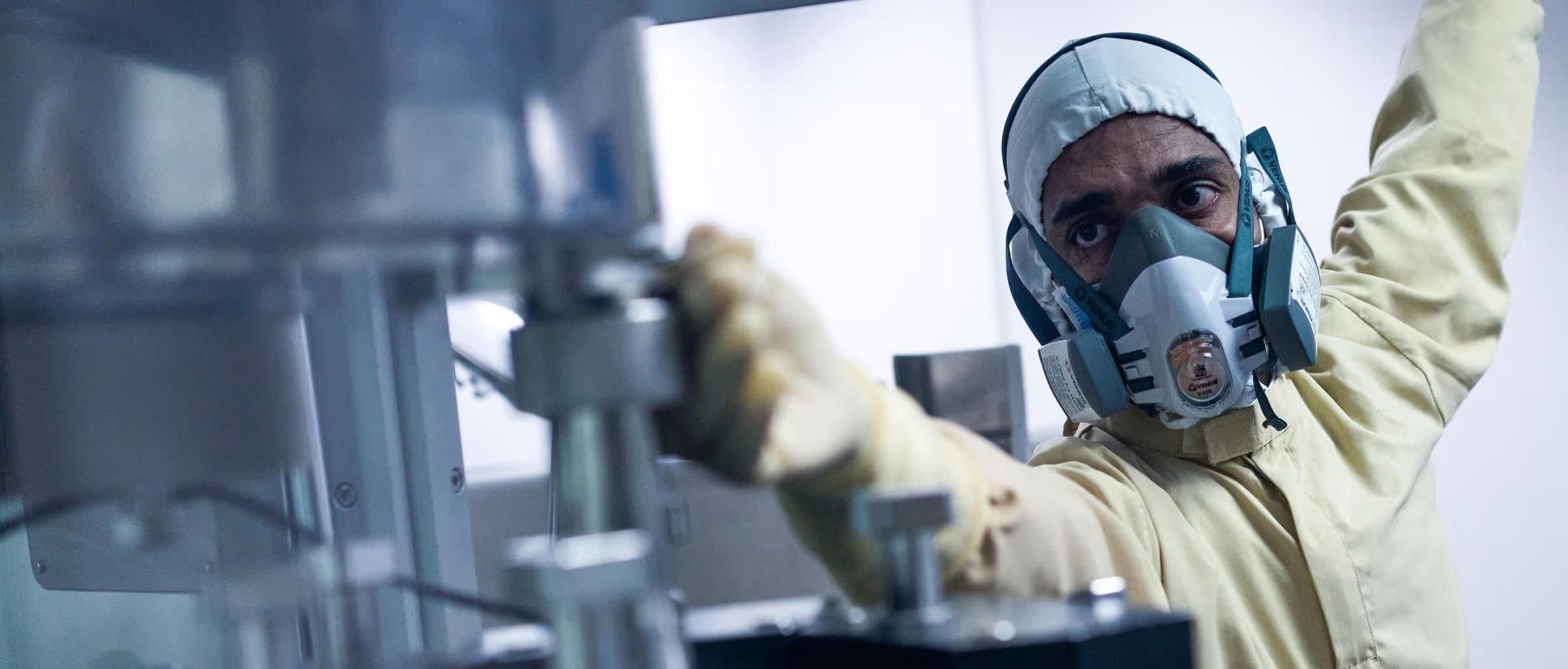 A man in a cream protective suit and gloves and heavy filtration mask operating a steel machine blurred in the foreground, depicting encapsulation of products at the capsule filling area, Goa, India