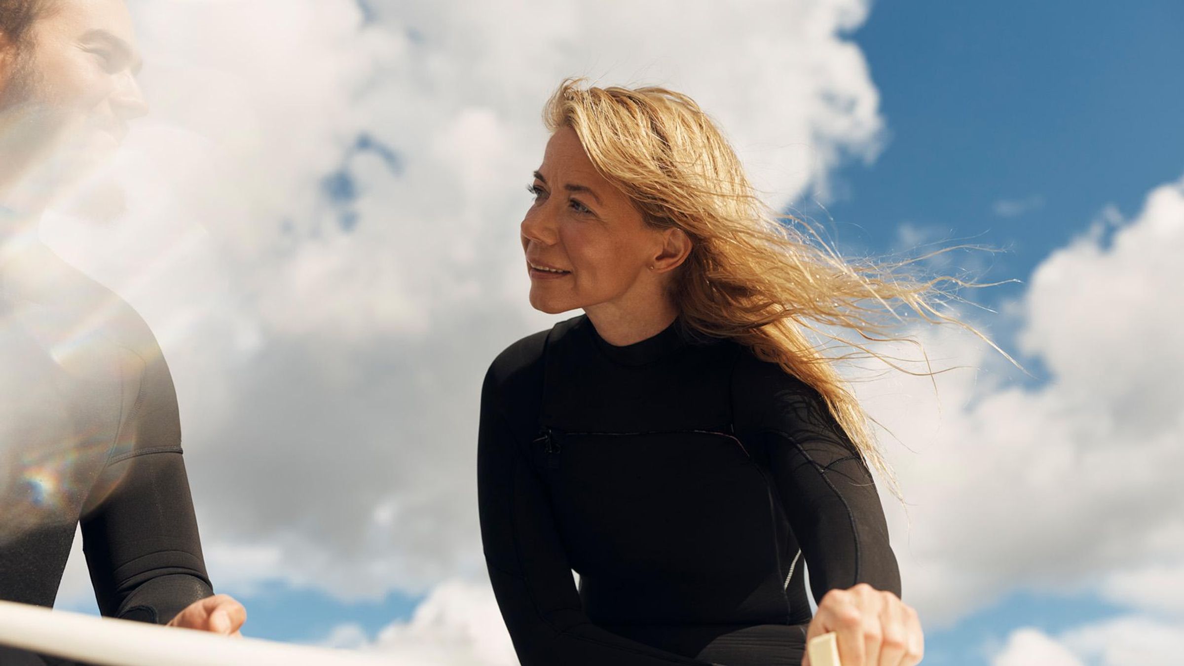 A man and a woman wearing black wetsuits look at each other happily while applying wax to a surfboard in the sunshine, with clouds and blue skies in the background.