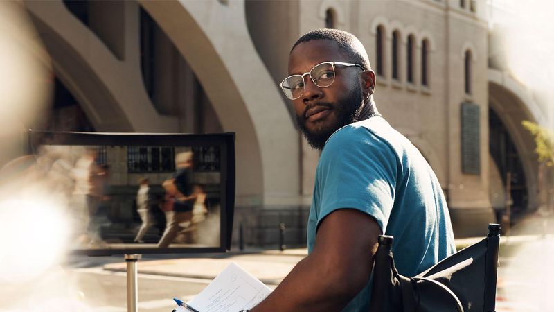 A man wearing glasses and a blue t-shirt sits in a director's chair and looks over his shoulder in the sunshine. He holds papers and a pen. In the background, a large monitor reflects blurred figures, and a bridge with arches is in the background.