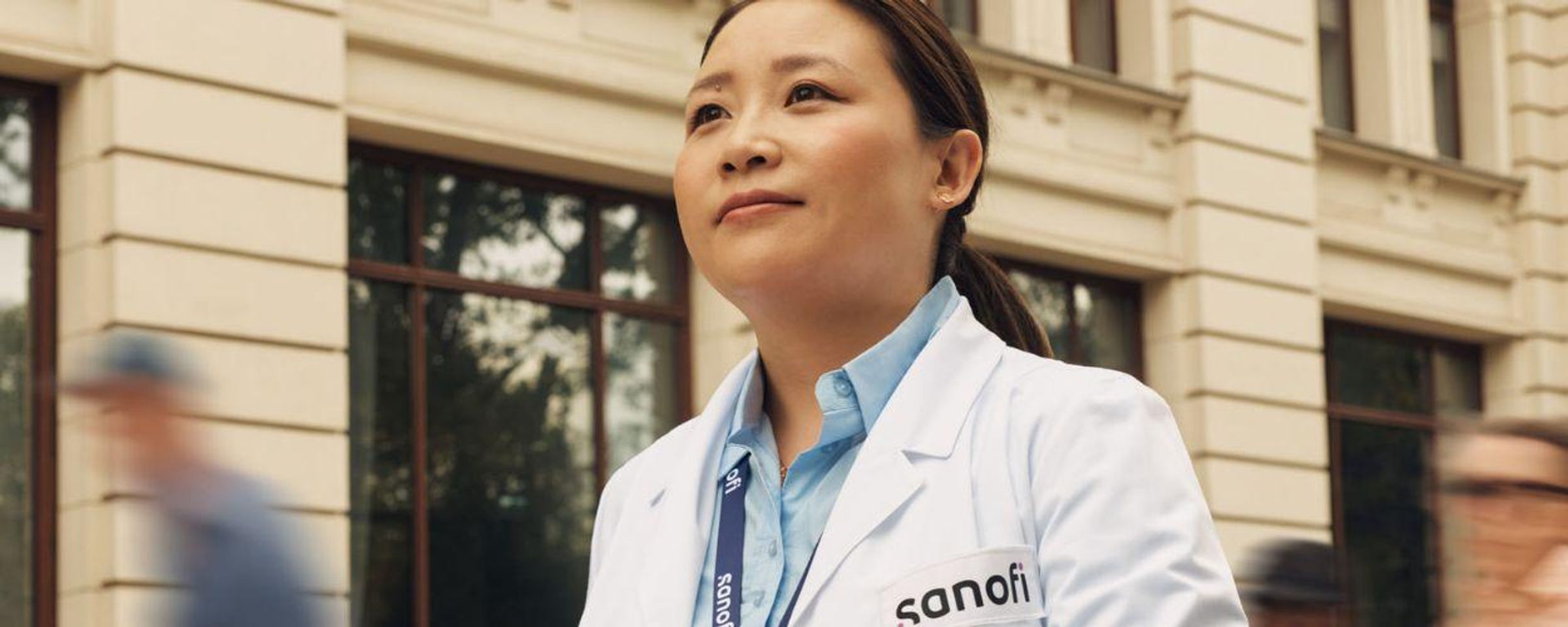 A woman in a white Sanofi lab coat and company badge outside of a building, moving forward while confidently and positively looking ahead. Several people are running alongside her in the background.