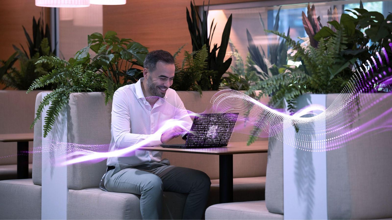 A person sitting in a modern, plant-filled workspace, working on a laptop at a booth-style table. Soft overhead lights illuminate the area, and a purple, wave-like digital graphic flows across the foreground.