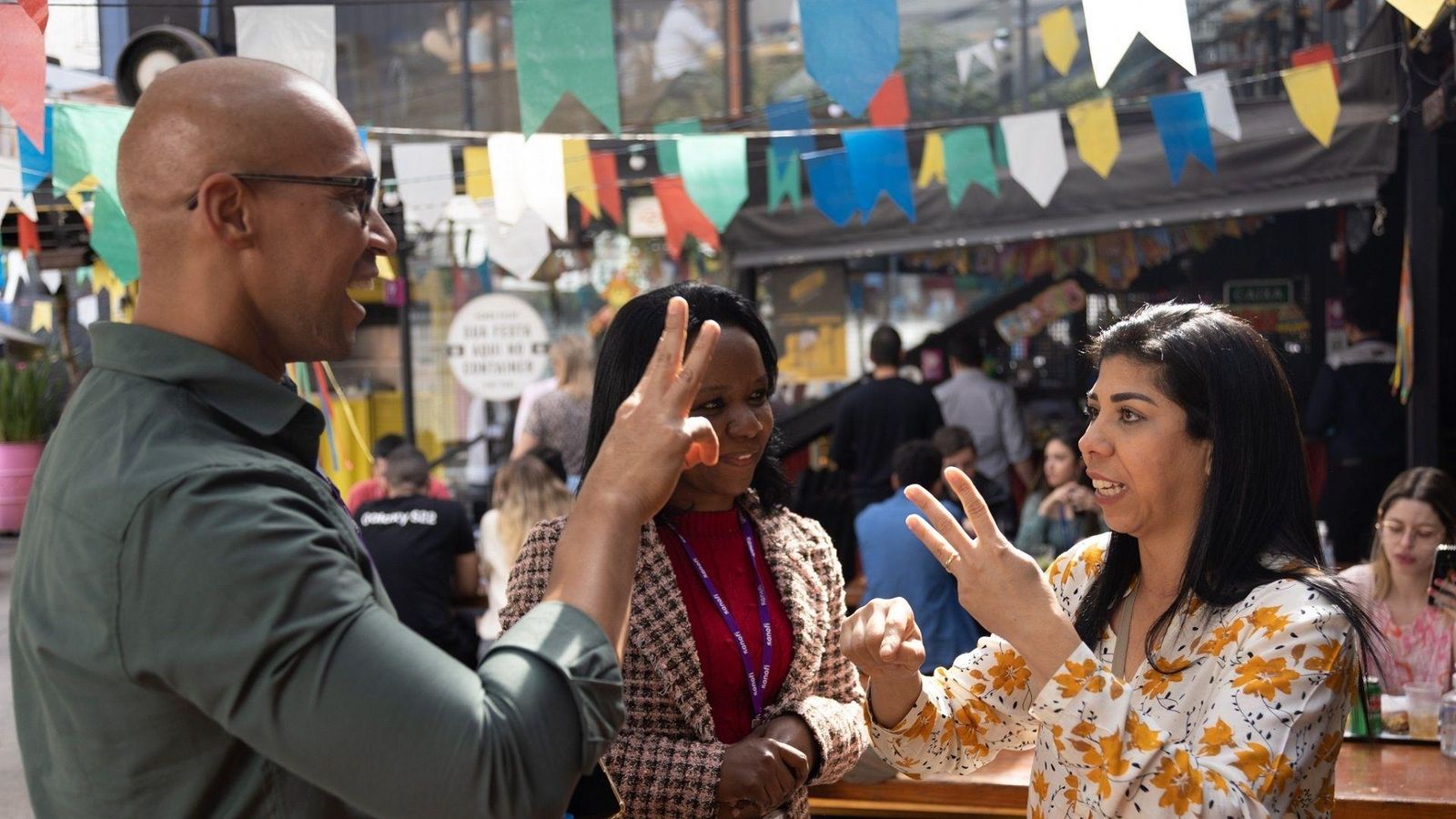 Three people conversing at a colorful outdoor market with festive decorations and bustling activity in the background.