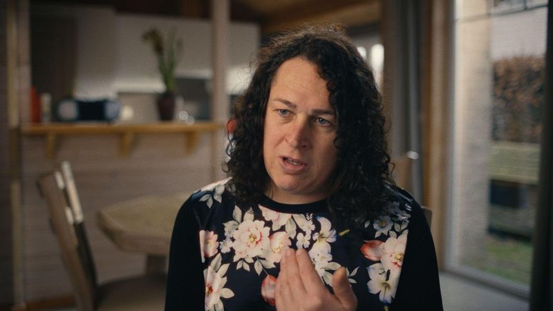 Jill Wuyts from Sanofi Belgium speaking expressively in a home setting, wearing a black floral top with a blurred living area in the background.