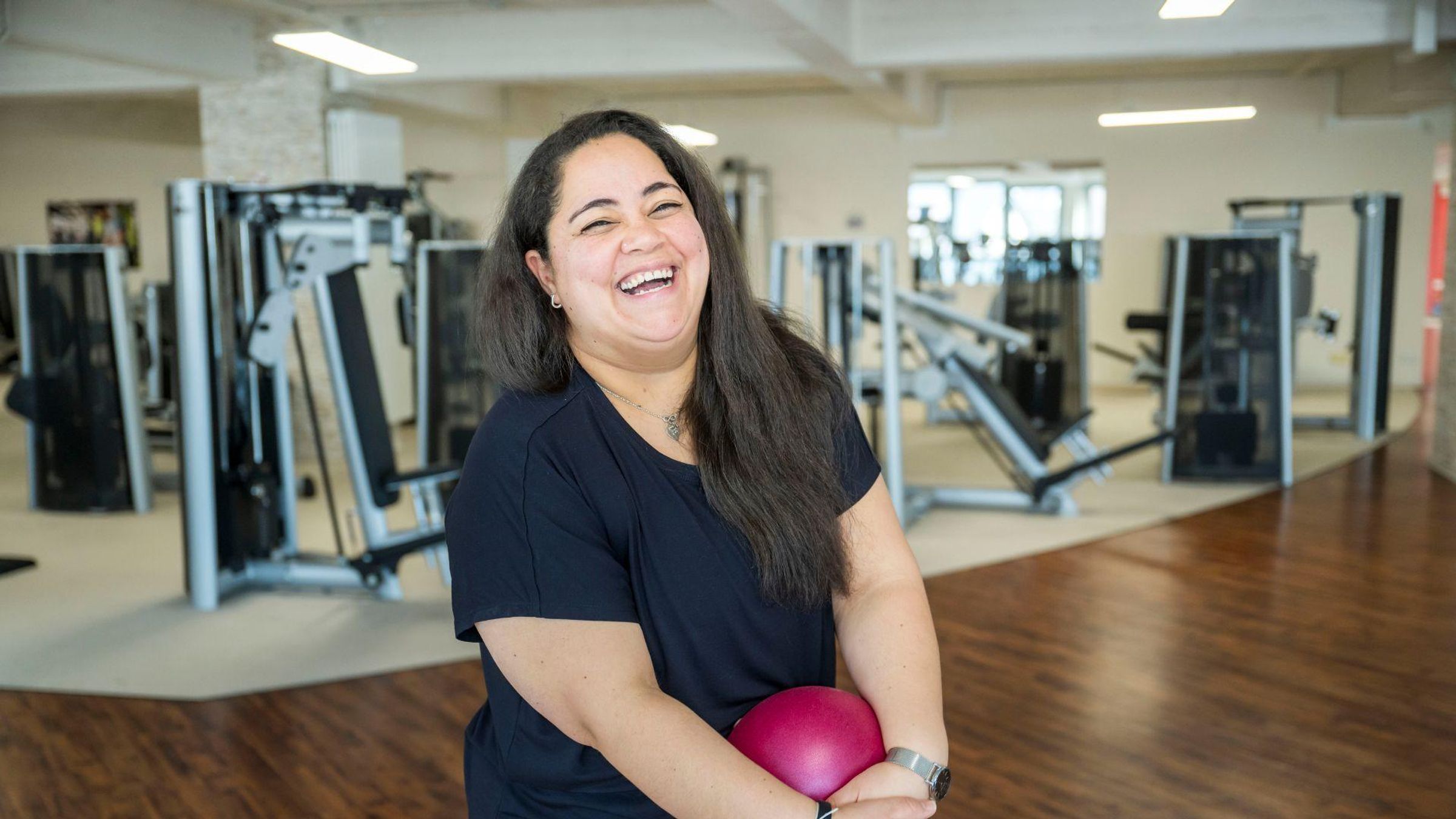 Woman standing in a gym holding a pink exercise ball.