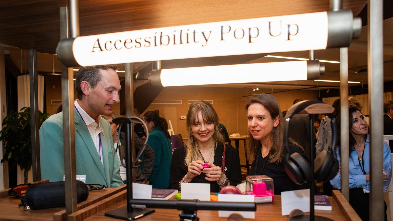A group of people gathered around an “Accessibility Pop Up” booth displaying assistive technology items, including headphones and tactile devices, inside a well lit event space.