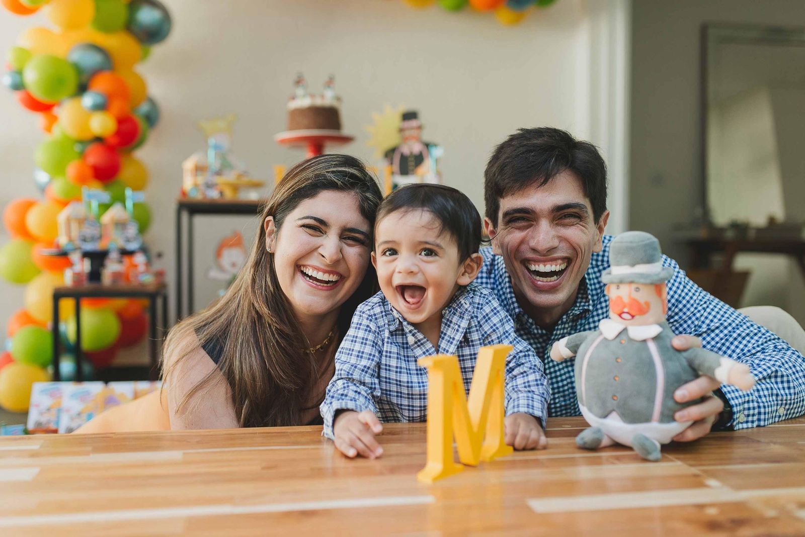 Smiling family - man, woman, and young boy in blue shirt - seated together at a wooden table with colorful balloons in the background