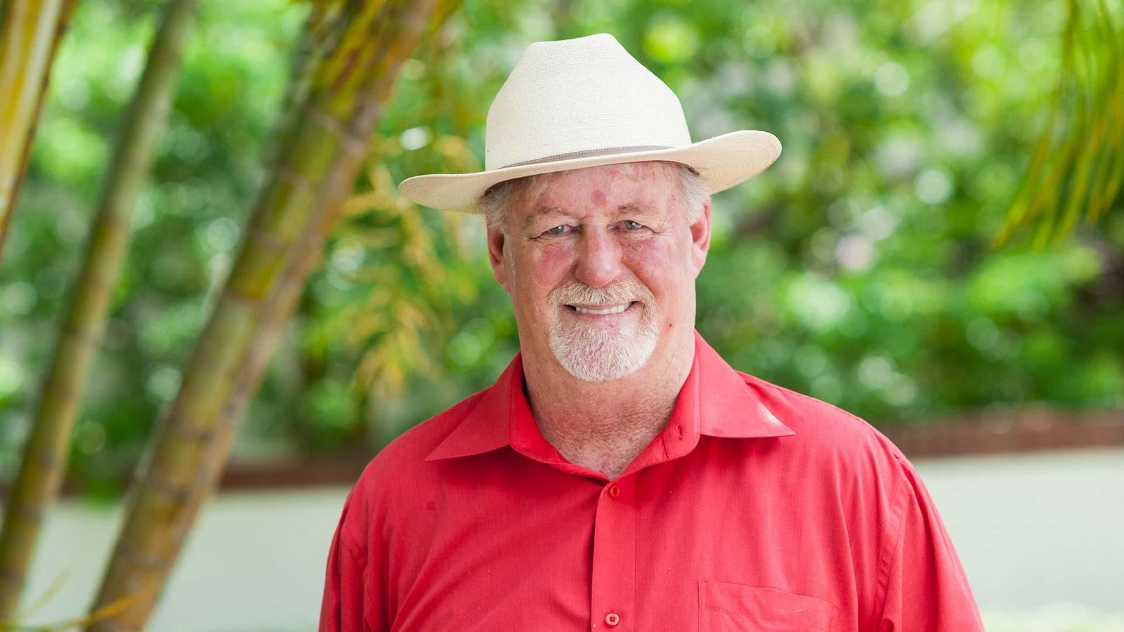 Portrait of Ron, an American patient with squamous cell carcinoma, smiling, wearing a white cowboy hat and red shirt, in a green outdoor setting