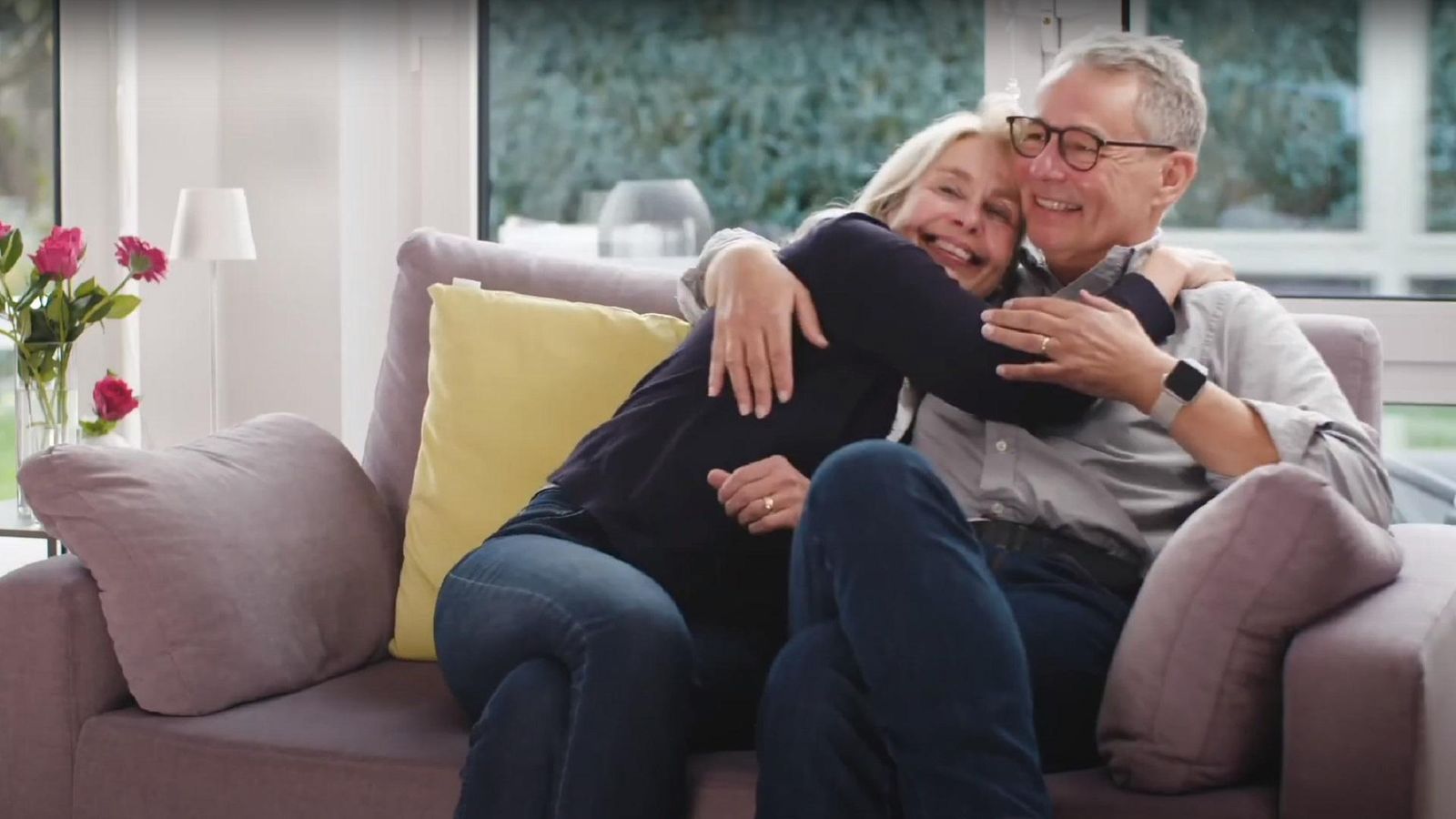 Elderly couple sitting on a couch in a living room, the man wearing glasses embraces the woman from behind, both looking off-camera