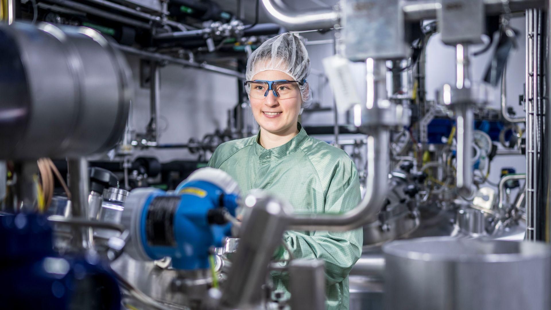 Julia Schwendner, Process Responsible Scientist, smiling in a green lab coat, hairnet and safety goggles while standing in front of industrial bioprocessing equipment at Sanofi's Frankfurt facility.