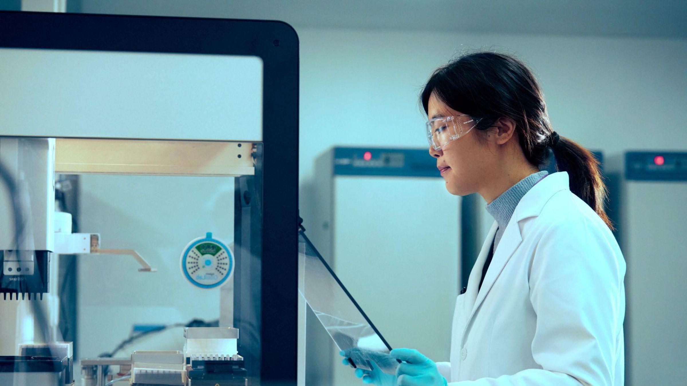 Shuang Yang works equipment in a glass case in a lab, wearing a white labcoat, safety glasses, and tied up dark hair