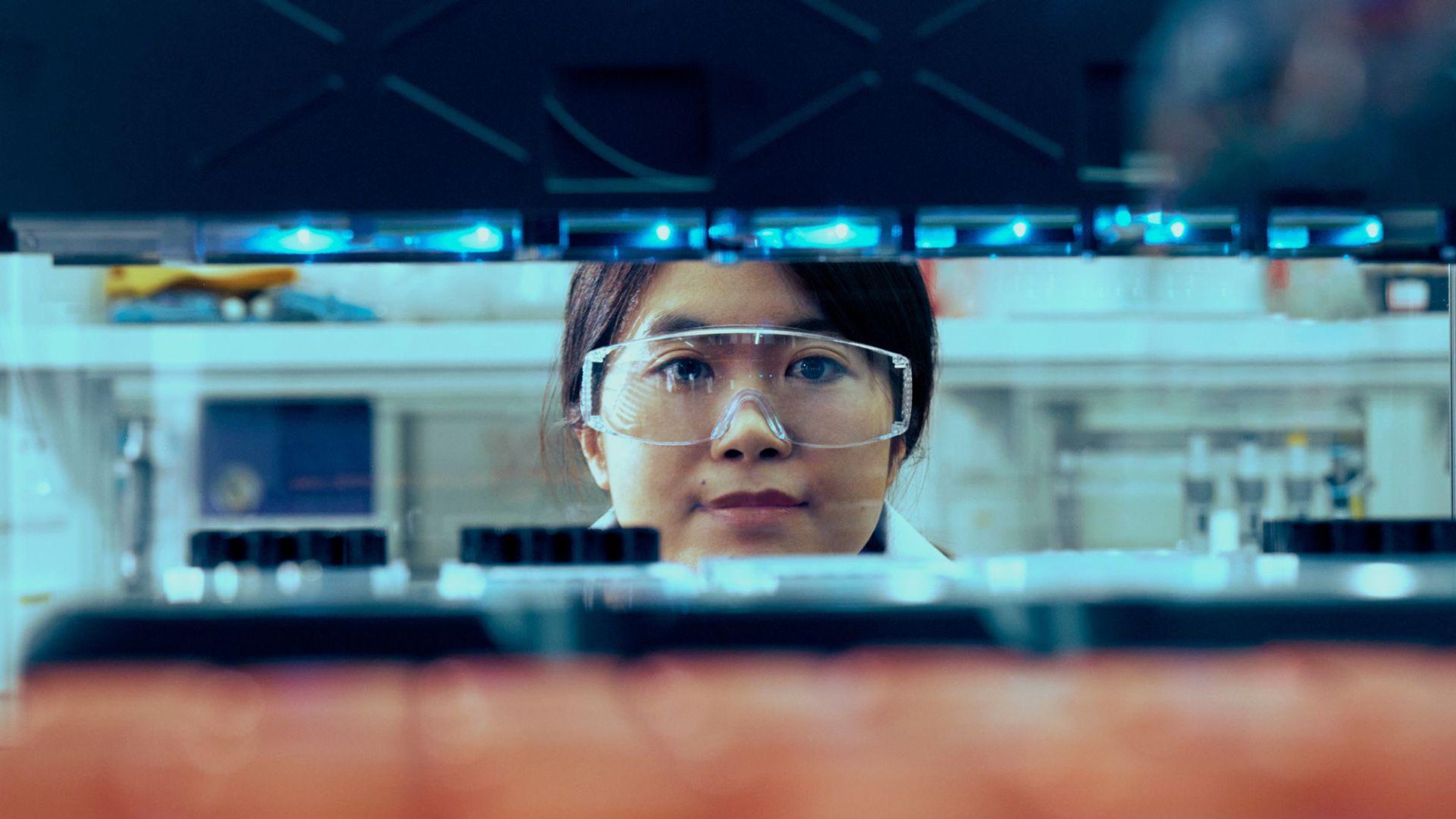 Shuang Yang, with dark tied up hair and safety glasses, as seen through some equipment in a lab, with only her head visible