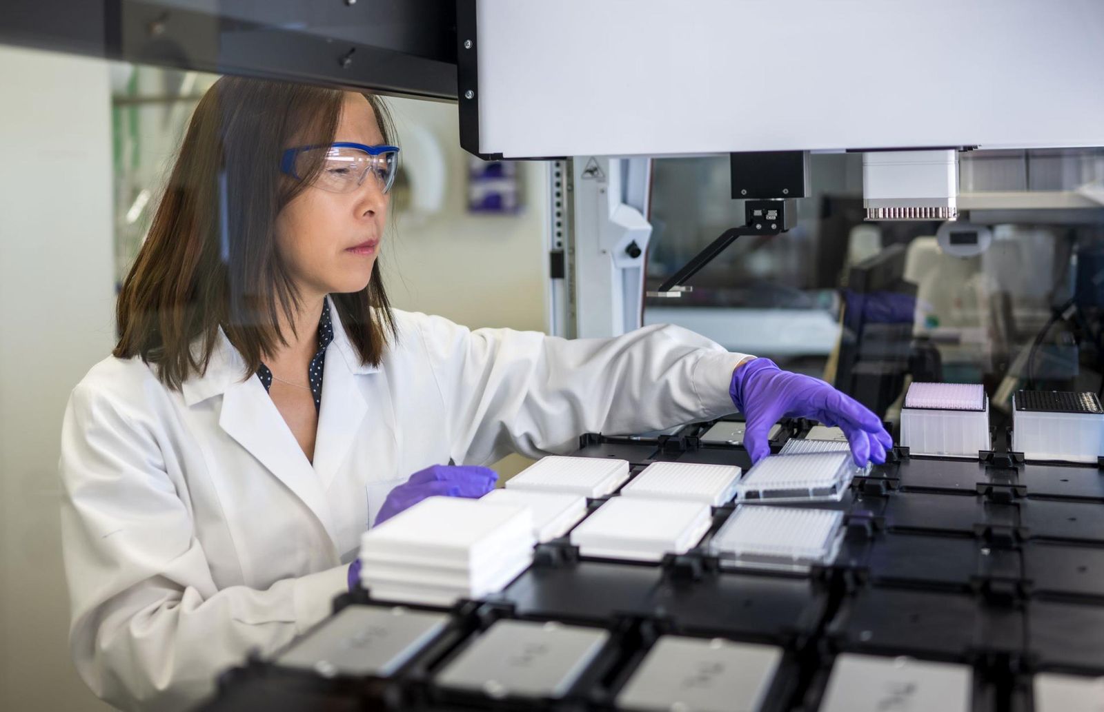 Ziyu Li inspects samples on a lab machine, wearing a white lab coat, purple gloves, and safety glasses.